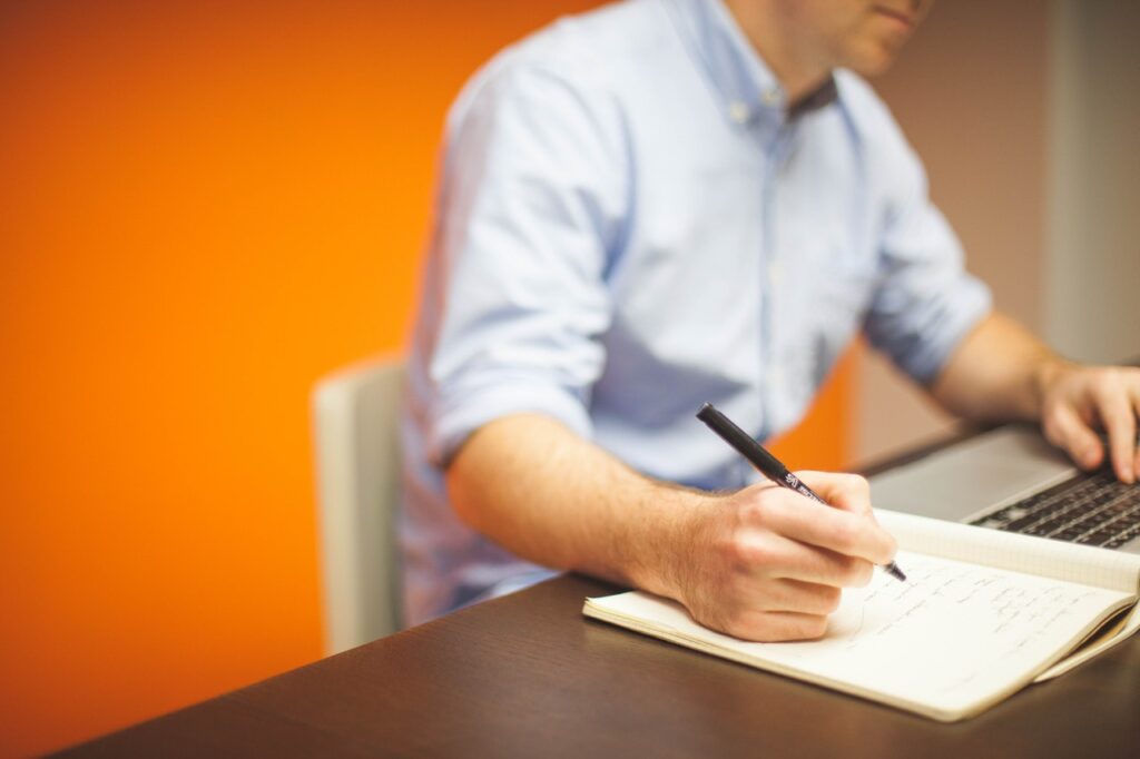 Person writing in notebook beside laptop.